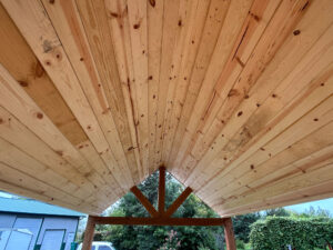 A perspective view from beneath the wooden roof of a carport or shelter, showcasing its natural timber planks and triangular truss structure. In the background, greenery and a neighboring building with a gray roof are visible.