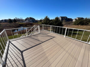 A sunlit outdoor deck with white railings overlooks a grassy yard and a small pond. Residential houses and evergreen trees are visible in the distance under a clear blue sky.