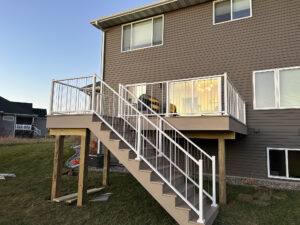 A light gray elevated deck with white railings and a staircase attached to a two-story brown house. The deck features patio furniture. The surrounding yard is grassy with neighboring houses visible in the background.