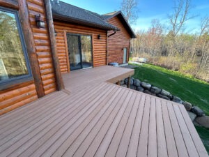 A view of a wooden deck attached to a log cabin-style house with sliding glass doors leading inside. The deck overlooks a green lawn bordered by rocks, with a wooded area in the background. The sky is clear and blue.