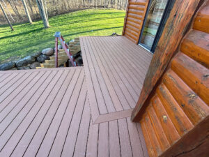 A wooden deck wraps around the corner of a log cabin, overlooking a grassy area with trees. A red ladder leans on the corner of the deck, and stacked stones are visible beside a wooden staircase under construction.