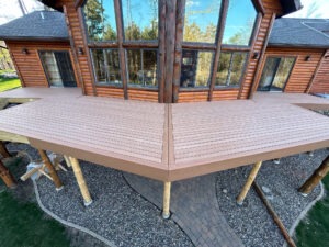 A view of a large wooden deck attached to a log cabin-style house. The deck is elevated on wooden pillars and has a geometric design with a central meeting point. Below is a stone pathway surrounded by gravel. Large windows and trees are visible in the background.